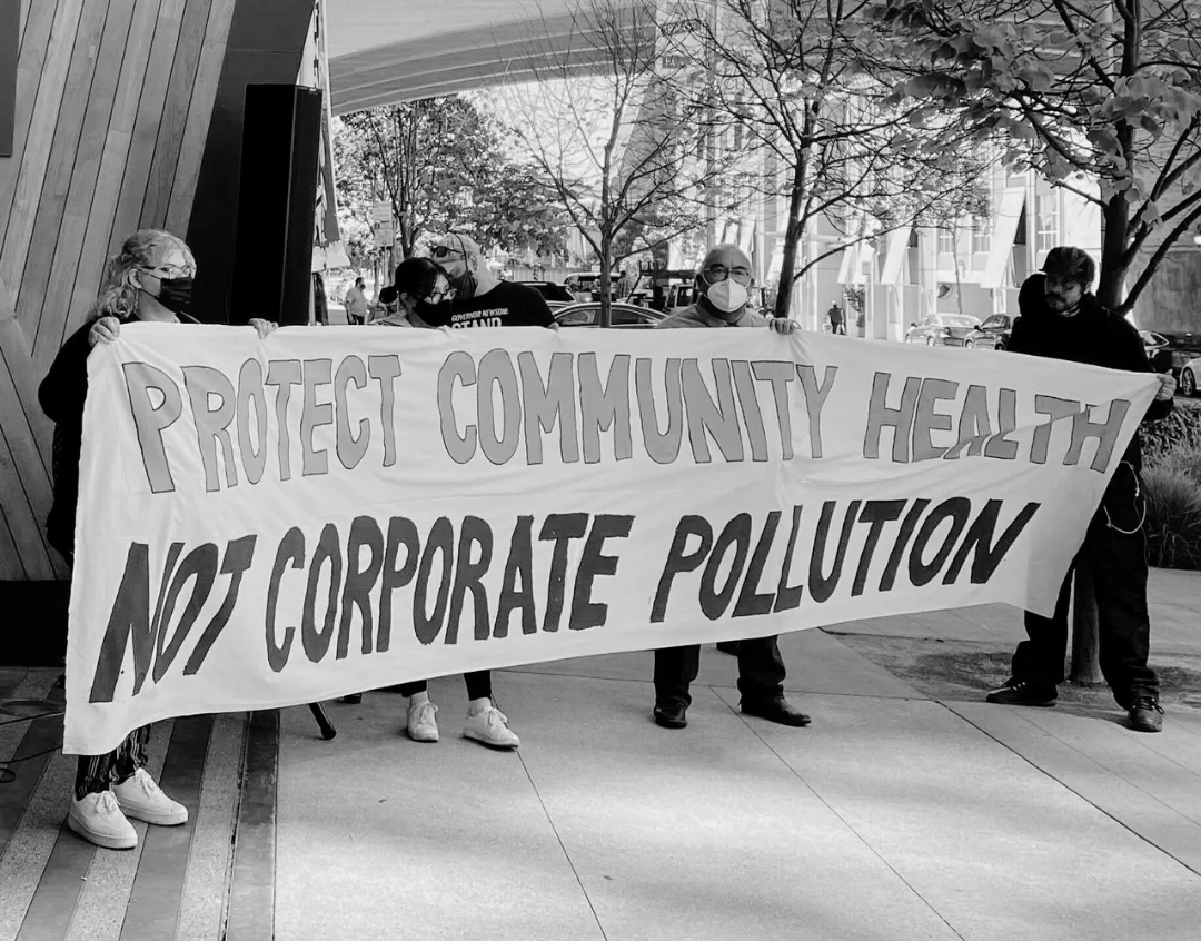 A group of protestors hold up a large banner that says Protect Community Health Not Corporate Pollution