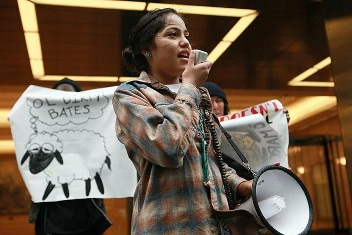 Young Native American girl speaking into megaphone at a 2018 protest to protect land and water rights. She stands in front of a large white banner with a big corporate building in the background representing corporate interests. The protest takes place in New York City.